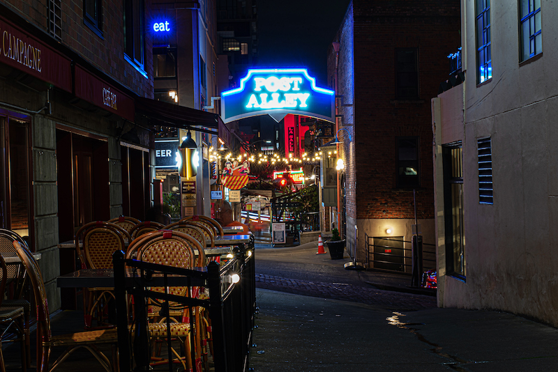 Dark alleyway at night leading to illuminated blue sign of Post Alley