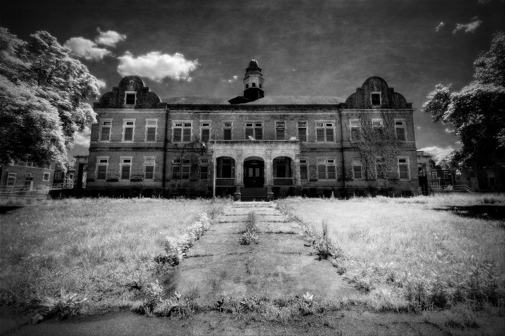 photo shows the facade of pennhurst in black and white, the grassy overgrown walkways leads up to the front doors and porch.