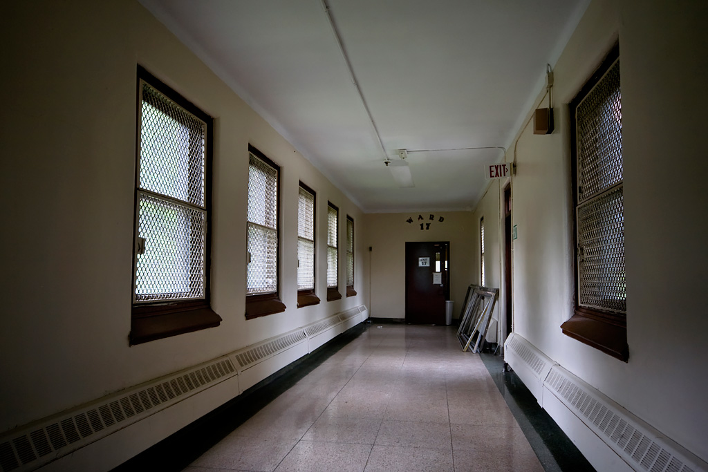 Photo shows a hospital hallway with a few old chairs and light pouring in through windows.