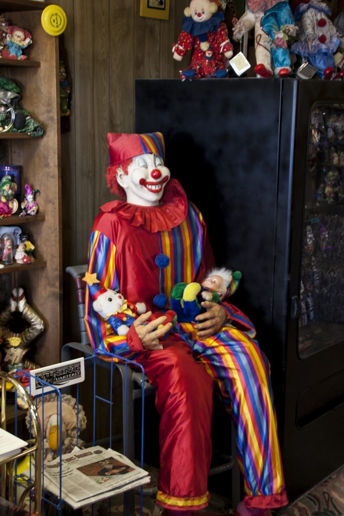 photo shows a life size clown sitting in front of a snack machine. 