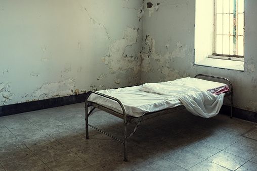 Abandoned bed in Mental Hospital, peeling paint on walls, filthy floors. Bed has sheets on it that look well used.