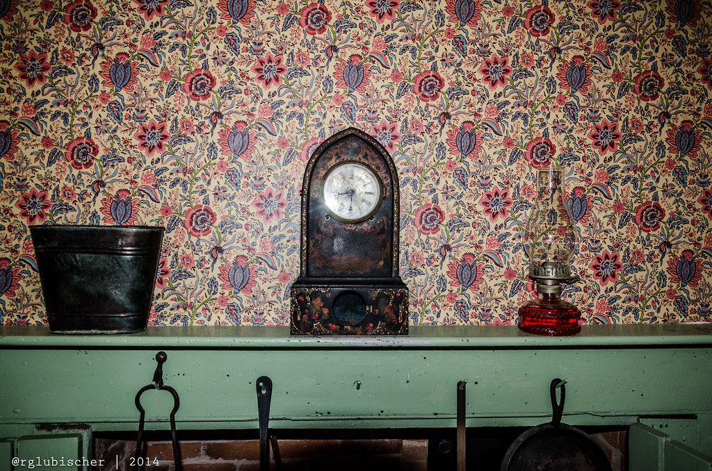 photo shows an old timepiece sitting on a mantle next to an oil lamp with floral wallpaper in the background