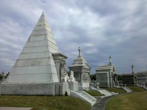 Metairie Cemetery. A pyramid shaped above ground tomb made from marble. Behind neo classical style tombs