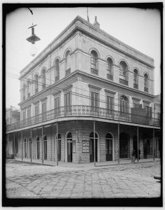Black and white photo of a large three story corner mansion with a balcony lining its exterior