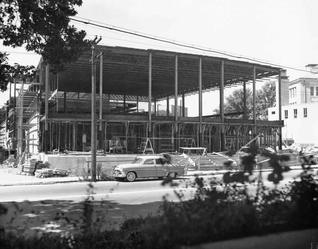 A black and white photo; the construction of a theatre in the 1960's. A classic car sits out front