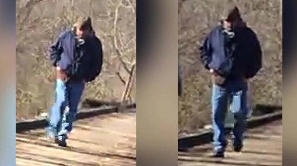 The Delphi Murders. Man in blue jeans and dark blue jacket and hat walking on wooden bridge. 