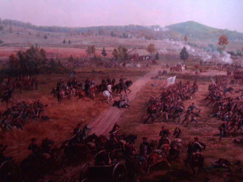 Paiting depicting the battle of Gettysburg. Soliders, horses and canons line eithe side of a road running through a series of hills. Smoke fills the air