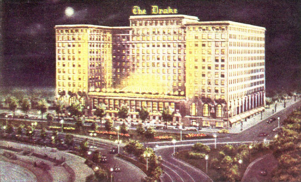 A large hotel lit up in front of a bending road. A lit up sign reading The Drake sits atop the building 