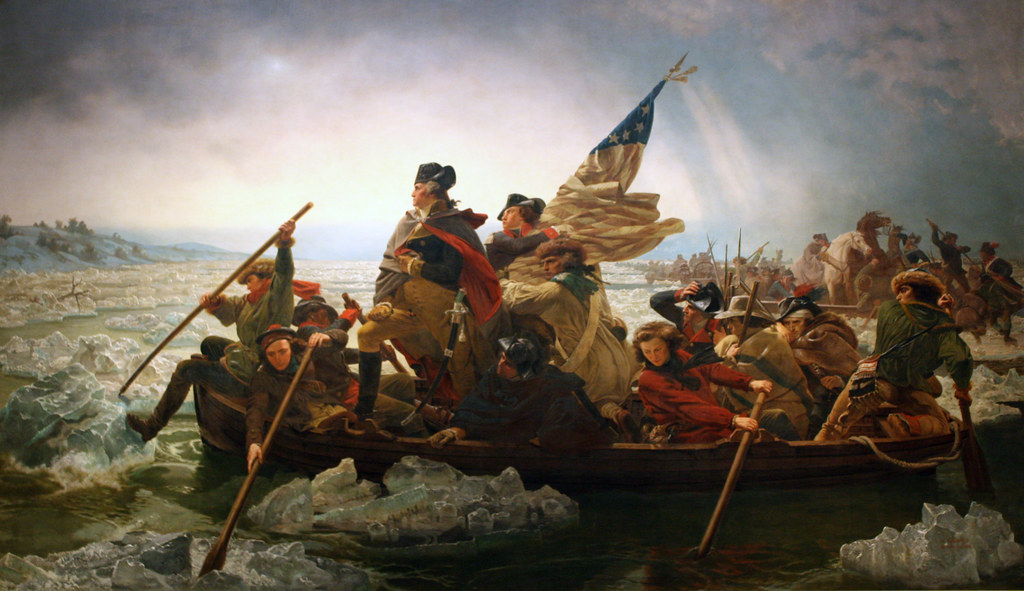 A painting of twelve men in a small boat paddling across treacherous water. At the front of the boat George Washington stands, one knee forward. An American flag waves above his head