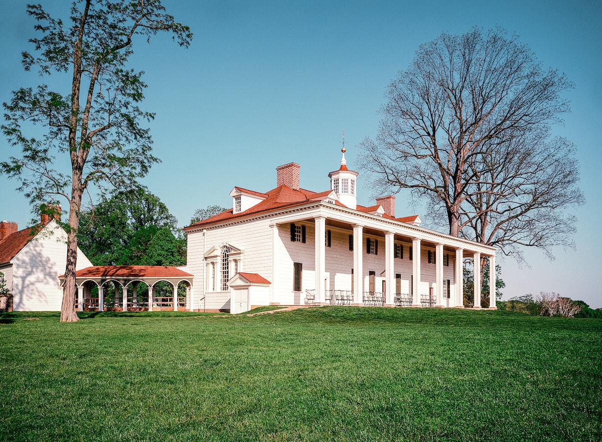 Photograph of a large plantation with red roof. Three barren trees stand in front of a clear blue sky. Green grass fills out the bottom of the photo.