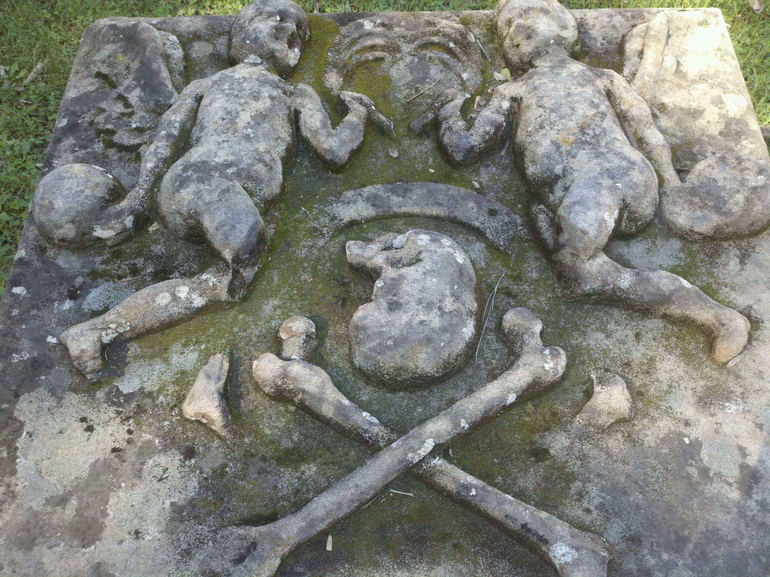 A skull and cross bones with two angles on either side adorn a tombstone covered in moss