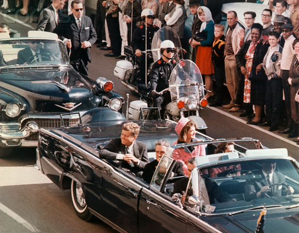 An open-topped car with John F. Kennedy and his wife, Jackie, in it driving down a street surrounded by a crowd