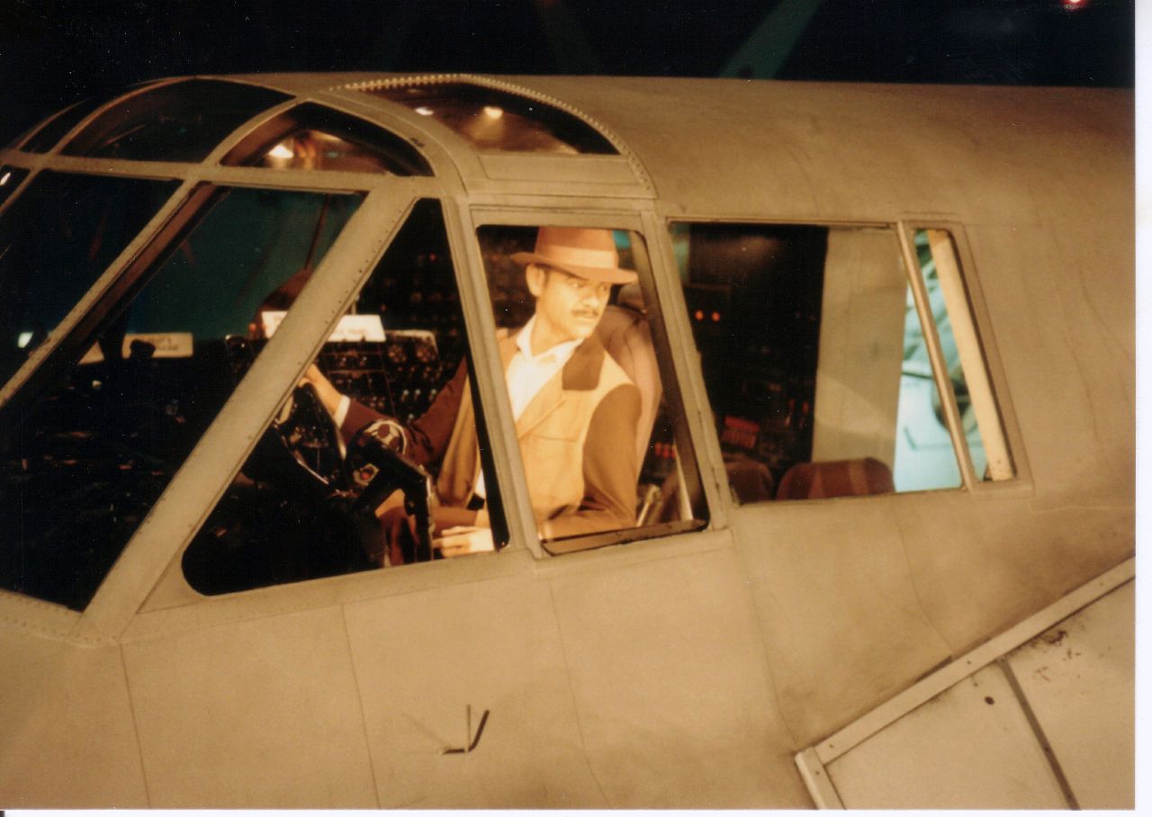 A man dressed in a brown suit with a brown hat leans out the window of a large plane