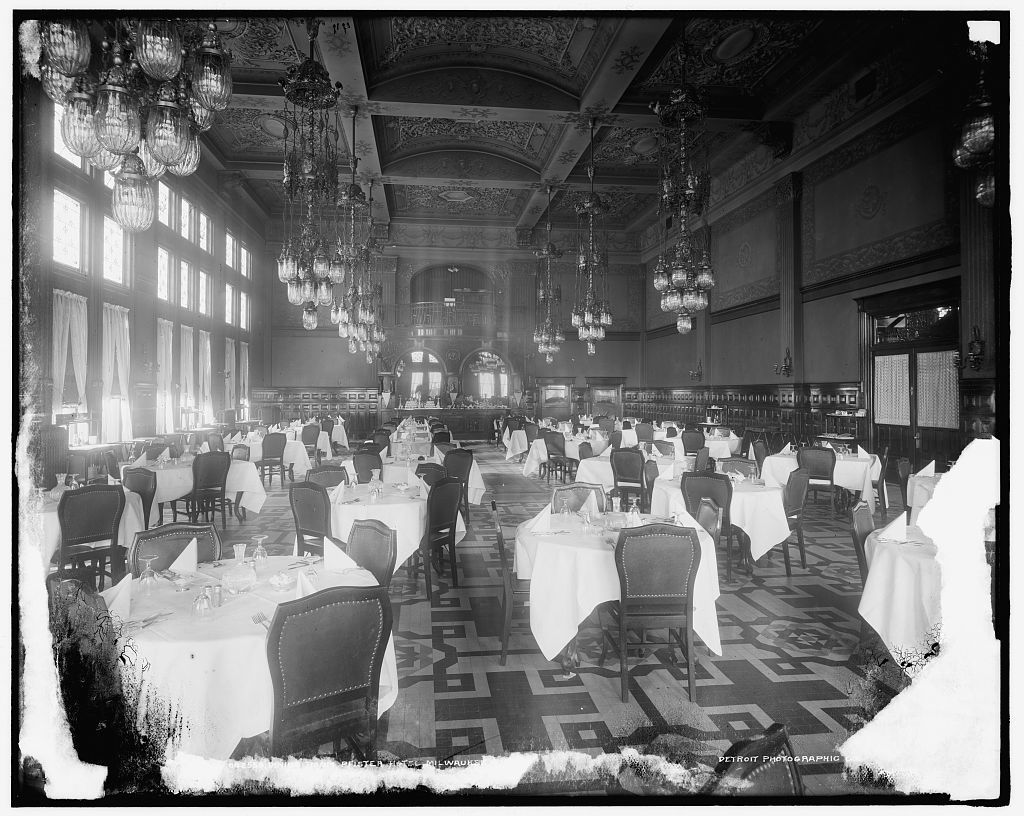 Black and white photo of a large dining room. High ceilings. It is full of people