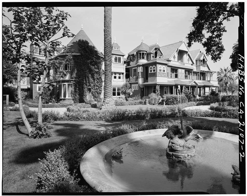 Black and white photo of a large mansion. A fountain and large palm tree are in the forefront