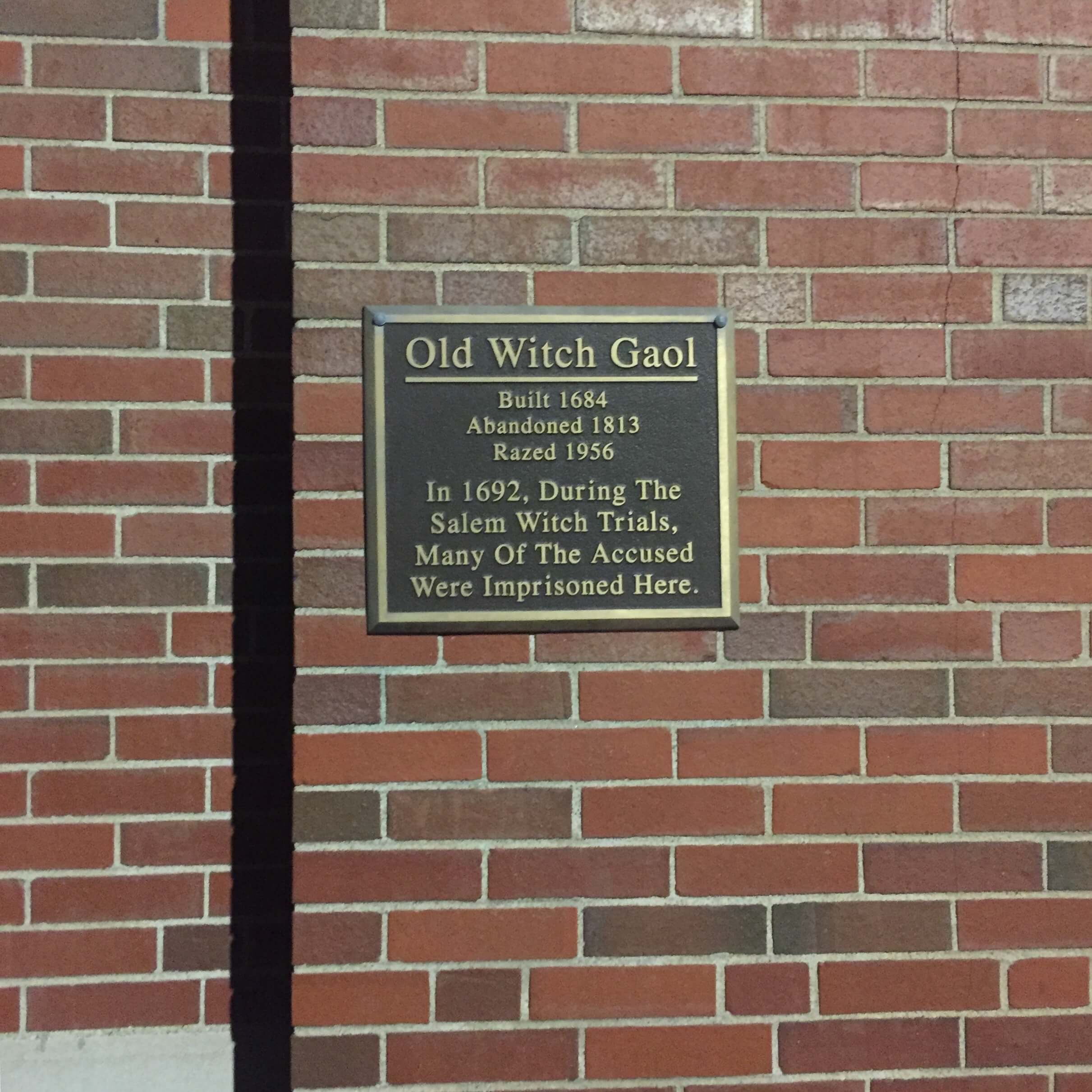 A plaque reading: Old Witch Goal: Built in 1684, Abandoned 1813, Razed 1956. In 1692, during the Salem Witch Trials, Many Of The Accused Were Imprisoned Here